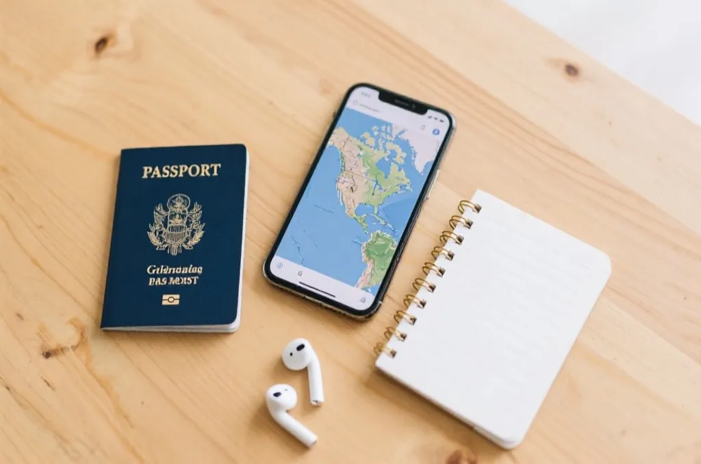 A flat lay of travel essentials on a clean wooden table: a generic passport, a smartphone showing a map of North America, a notebook, and wireless earbuds